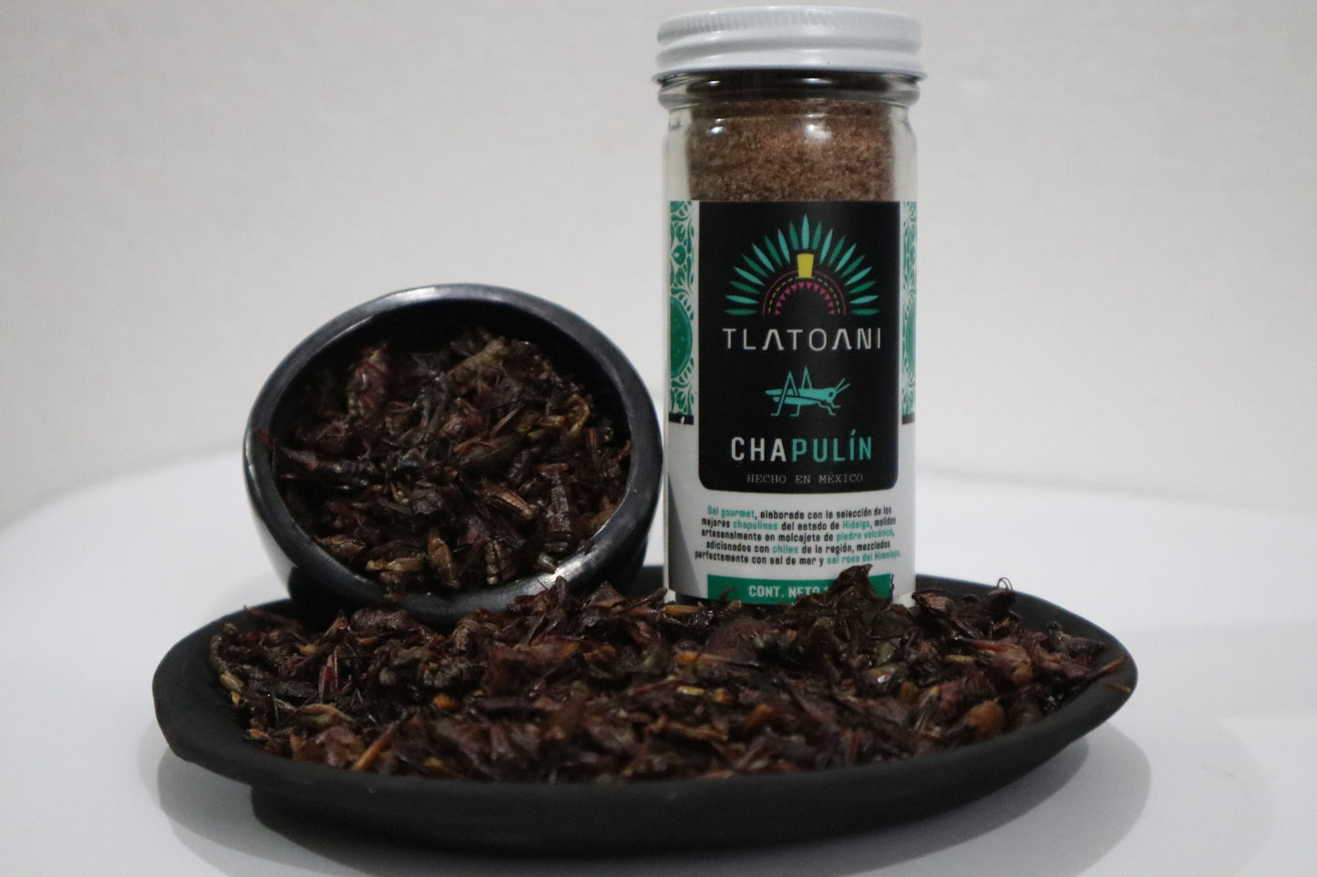 Frasco grande de sal de chapulín Tlatoani junto a platos de barro negro con chapulines enteros tostados, presentados sobre fondo blanco.