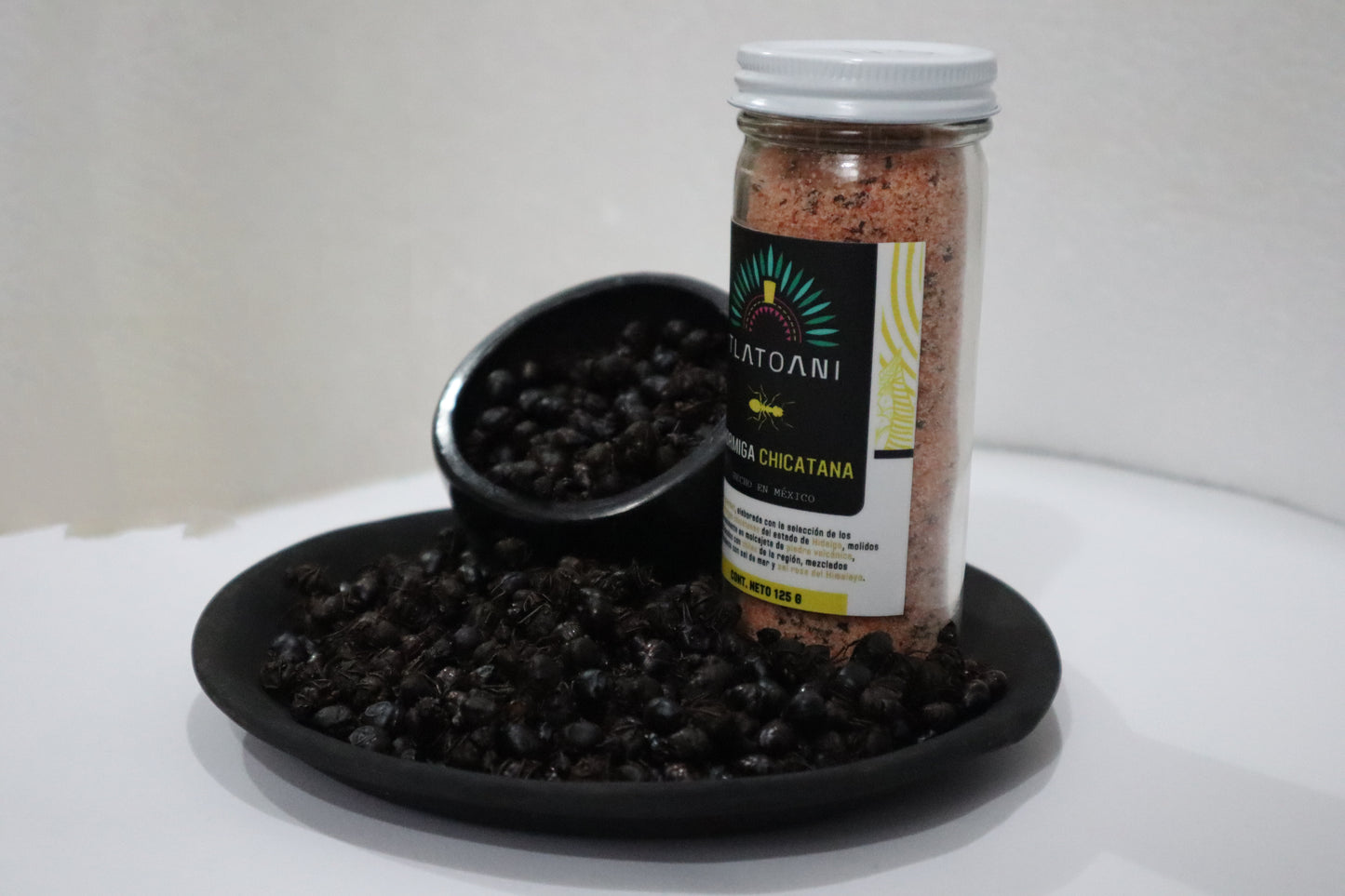Frasco grande de sal gourmet Tlatoani de hormiga chicatana sobre un plato negro lleno de hormigas enteras tostadas.