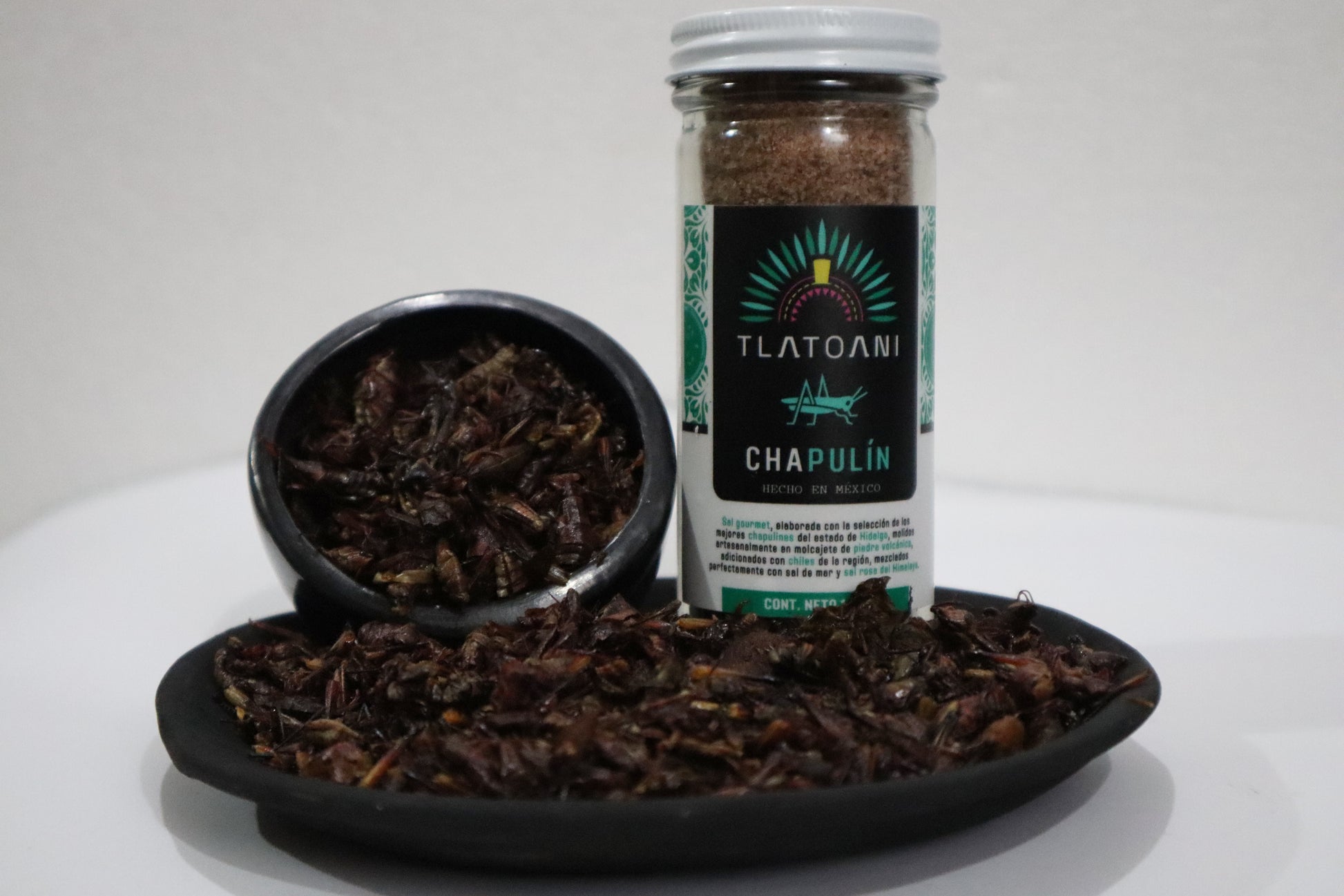 Frasco grande de sal de chapulín Tlatoani junto a platos de barro negro con chapulines enteros tostados, presentados sobre fondo blanco.