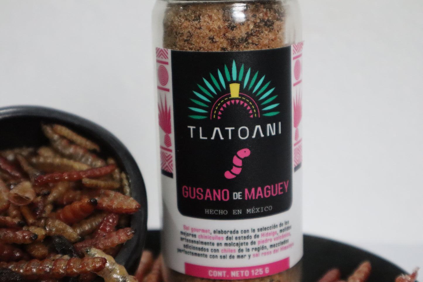 Frasco grande de sal de gusano de maguey marca Tlatoani, acompañado de un recipiente de barro con gusanos secos, ingredientes clave de esta sal tradicional mexicana.