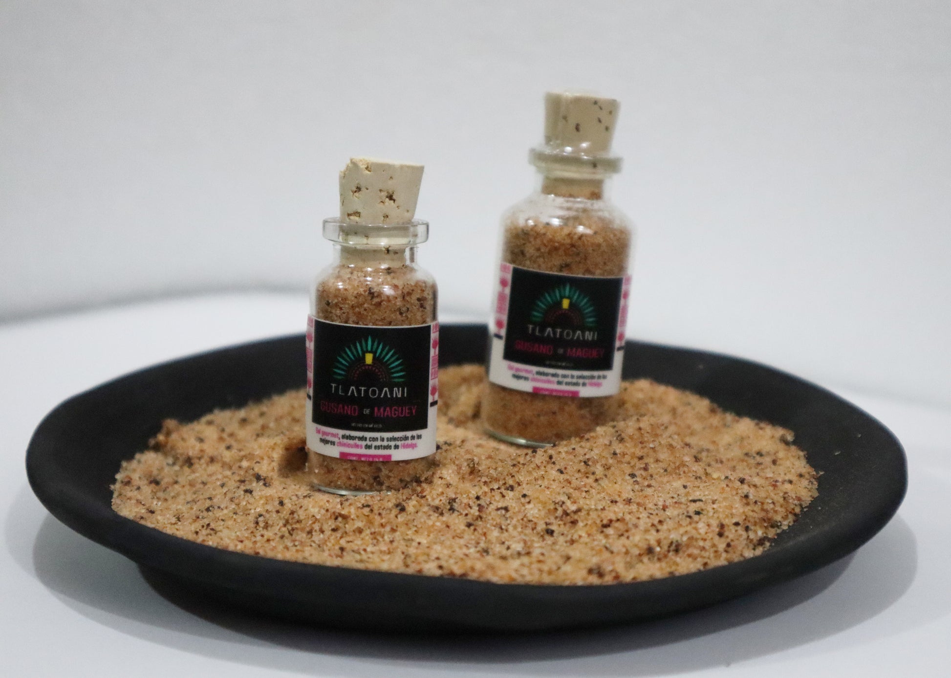 Dos frascos de sal de gusano de maguey marca Tlatoani, en presentación chica y mediana, colocados sobre un plato de barro lleno de la misma sal gourmet.