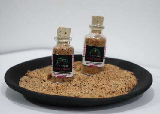 Dos frascos de sal de gusano de maguey marca Tlatoani, en presentación chica y mediana, colocados sobre un plato de barro lleno de la misma sal gourmet.