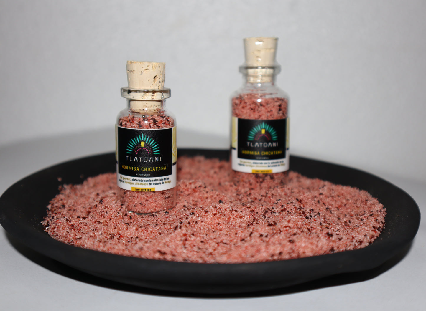 Frascos pequeños de sal gourmet Tlatoani de hormiga chicatana, colocados sobre una base de sal color rojiza con trozos visibles de hormiga.