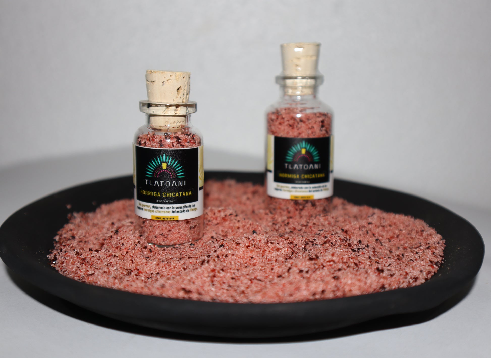 Frascos pequeños de sal gourmet Tlatoani de hormiga chicatana, colocados sobre una base de sal color rojiza con trozos visibles de hormiga.