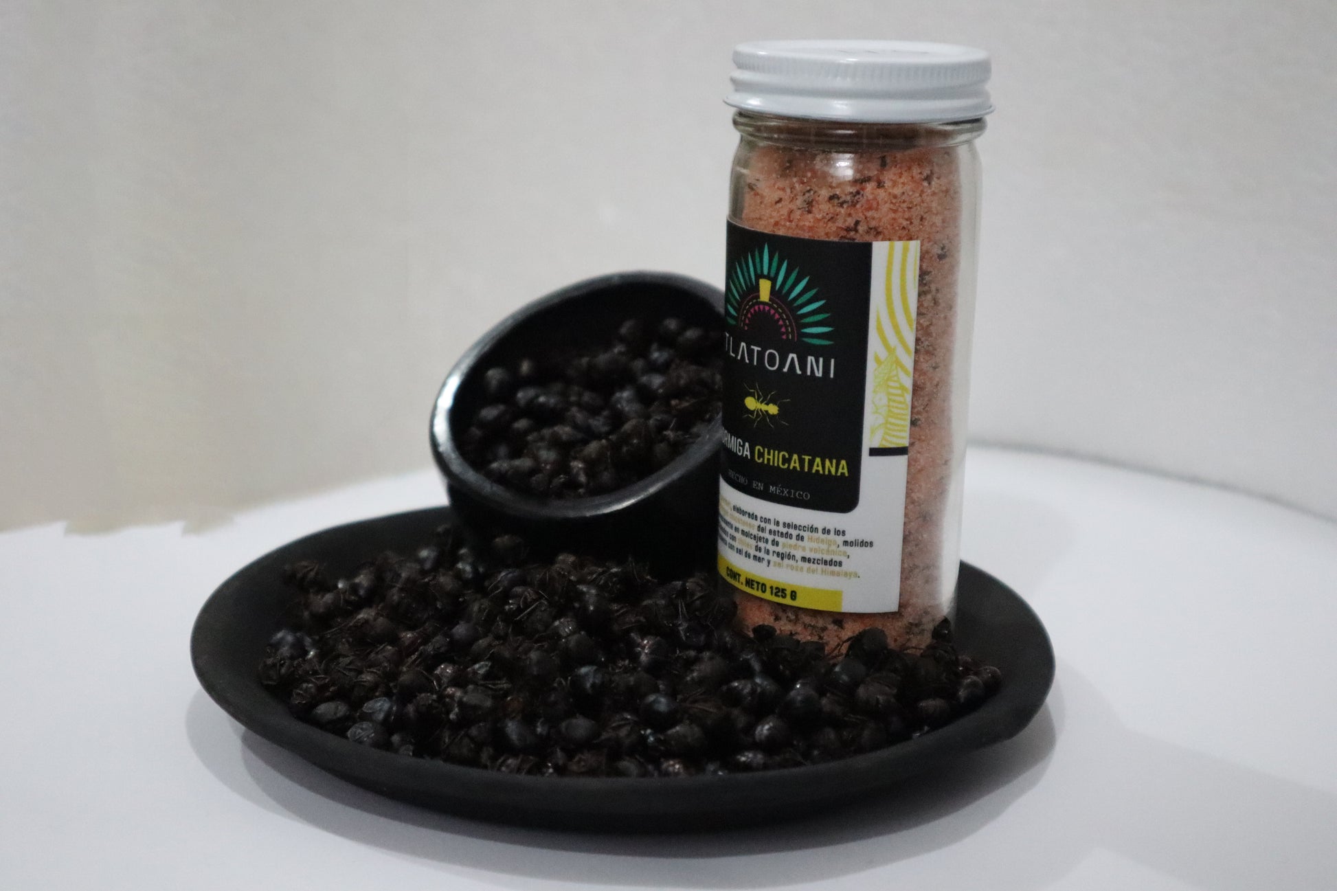 Frasco grande de sal gourmet Tlatoani de hormiga chicatana sobre un plato negro lleno de hormigas enteras tostadas.